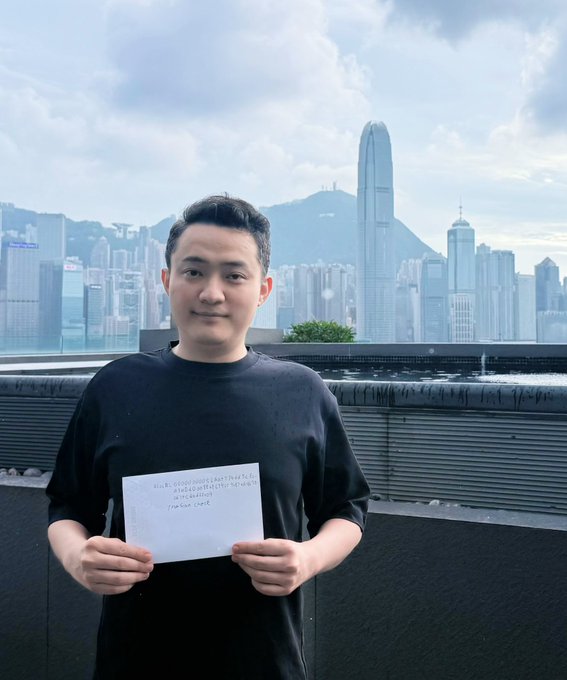 A young man in a black t-shirt holding a handwritten note in front of the Hong Kong skyline, with the iconic IFC tower visible in the background.
