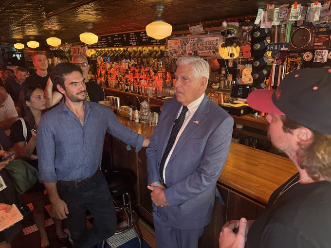 An older man in a gray suit speaks with a younger man in a blue shirt inside a bar filled with liquor bottles and sports memorabilia.