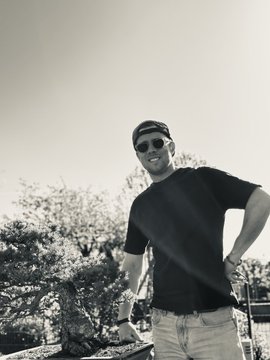 A black-and-white photo of a smiling man wearing sunglasses, a t-shirt, and a backwards baseball cap, standing outdoors with trees in the background.