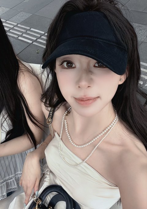 The image features a woman with long hair wearing a white dress, sitting down while holding a black purse. She has a hat on her head that matches her outfit. The background of the photo appears to be a gray sidewalk or street, which contrasts nicely with the woman's attire and adds depth to the scene.