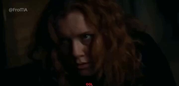 The image shows a close-up of a woman with long red hair, looking down at her hands while wearing a black jacket. The background appears to be dark, which contrasts with the woman's vibrant appearance. There are no texts or other objects in the image, and the focus is solely on the woman and her expression.