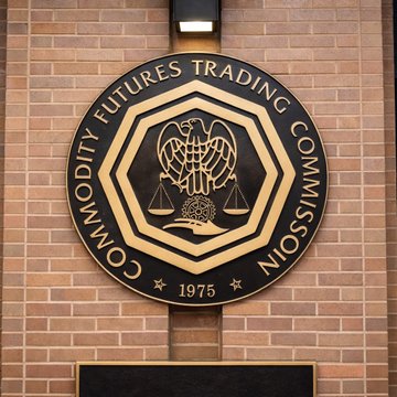 A circular bronze and gold seal of the **Commodity Futures Trading Commission (CFTC)**, dated 1975, hangs on a brick wall. The seal relates directly to the CFTC's staff advisory on "mention market" contracts, as this is the regulatory body issuing the warning about heightened manipulation risks.