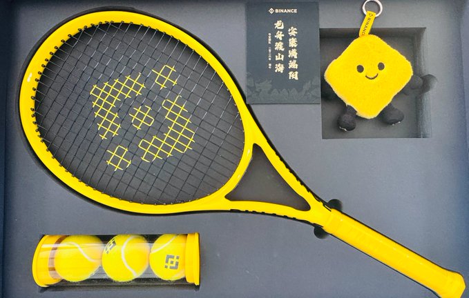 The image features a yellow tennis racket with a black handle, placed inside a box along with other tennis balls. The tennis ball packaging includes a small square-shaped toy that resembles a smiling face. The background of the image is dark gray, which contrasts with the bright colors of the tennis equipment and adds depth to the scene.