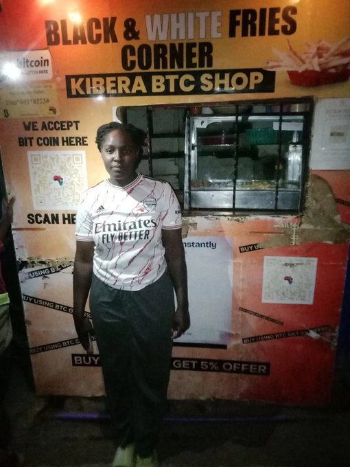 A woman in a white 'Emirates Fly Better' soccer jersey stands in front of a 'BLACK & WHITE FRIES CORNER' and 'KIBERA BTC SHOP' storefront that advertises Bitcoin acceptance and offers a 5% discount for using BTC.