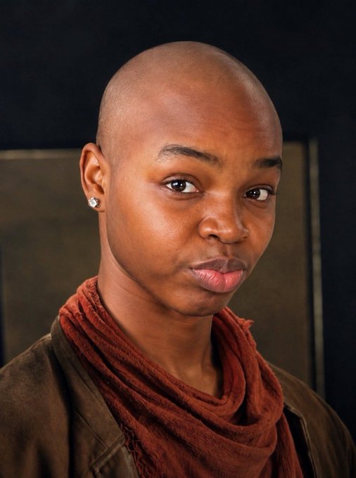 A close-up portrait of a bald person with a serious expression, wearing a red scarf and stud earrings.