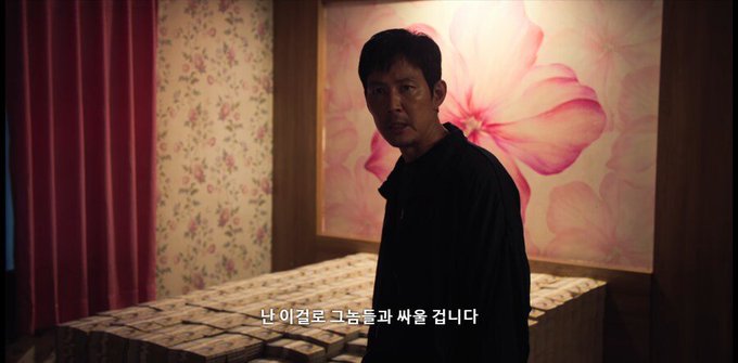 A man in a dark jacket stands in a dimly lit room with floral wallpaper and a large pink flower painting, looking over his shoulder at what appears to be stacks of money on a table. The Korean subtitle reads "난 이걸로 그놈들과 싸울 겁니다" ("I will fight those bastards with this"), reinforcing the post's theme of using SPCX funds aggressively to battle against others.