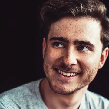 A close-up portrait of a smiling young man with brown hair, freckles, and light stubble, looking off to the side.