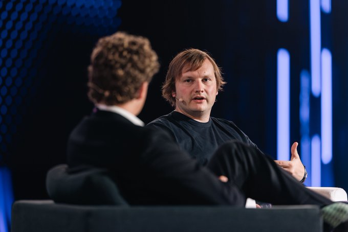 Two men sit in chairs on a stage during what appears to be a panel discussion or interview, with blue-lit vertical panels in the background.