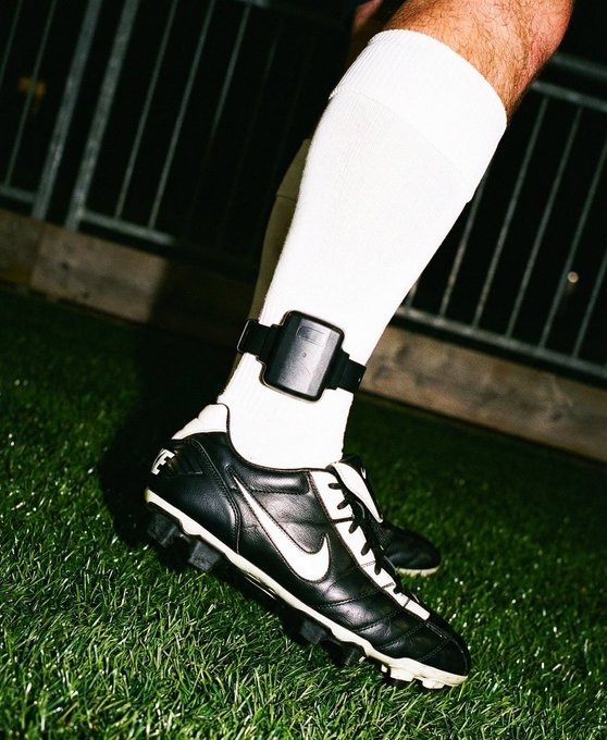 Close-up of a person's lower legs wearing white athletic socks with an ankle monitor strapped around one ankle, paired with black and white Nike soccer cleats on artificial turf.