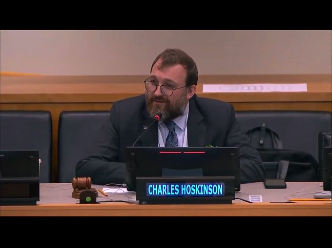 A photograph of Charles Hoskinson, founder of Cardano, seated at a panel table with a digital nameplate reading 'CHARLES HOSKINSON', with a wooden gavel visible on the desk.