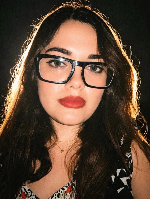A close-up portrait of a woman with dark hair, black-framed glasses, red lipstick, and a delicate gold necklace.