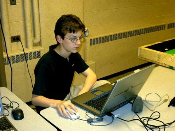 A young man in a black polo shirt and glasses sits at a desk working intently on a laptop, with cables, a mouse, and equipment scattered around him in what appears to be a lab or classroom setting. The image visually represents the post's theme of focused, systematic analysis—mirroring the deliberate, disciplined approach to compounding capital across $BTC, $ETH, and $SOL with explicit risk limits rather than frequent trading.