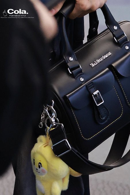 A person holds a black Dr. Martens handbag with a small yellow plush keychain, with a 'Cola.' logo in the corner.