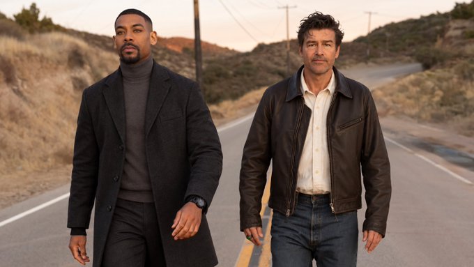 Two men stand on a desert road, one in a dark coat and turtleneck, the other in a brown leather jacket and light shirt.