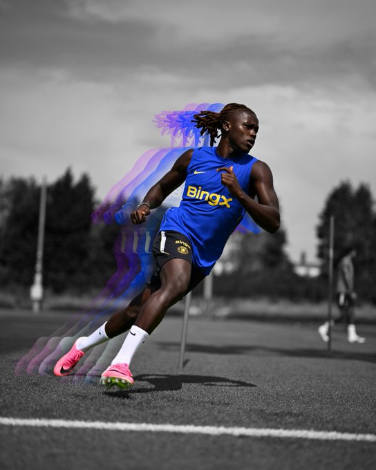 A Chelsea FC player sprints on a training pitch with a purple motion-trail effect highlighting his speed, wearing a blue kit with "BingX" sponsor text on the chest and matching BingX-branded shorts, paired with bright pink cleats that pop against the desaturated background—capturing the rapid, high-energy training moment referenced by the post's "blink and you'll miss them" tagline and #TrainedOnGreatness #BingXCFC campaign promoting the Bing x Chelsea FC collaboration.