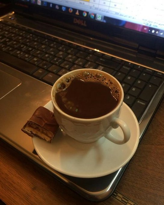 A small white cup of dark coffee sits on a saucer next to a piece of chocolate, placed on top of a Dell laptop keyboard.
