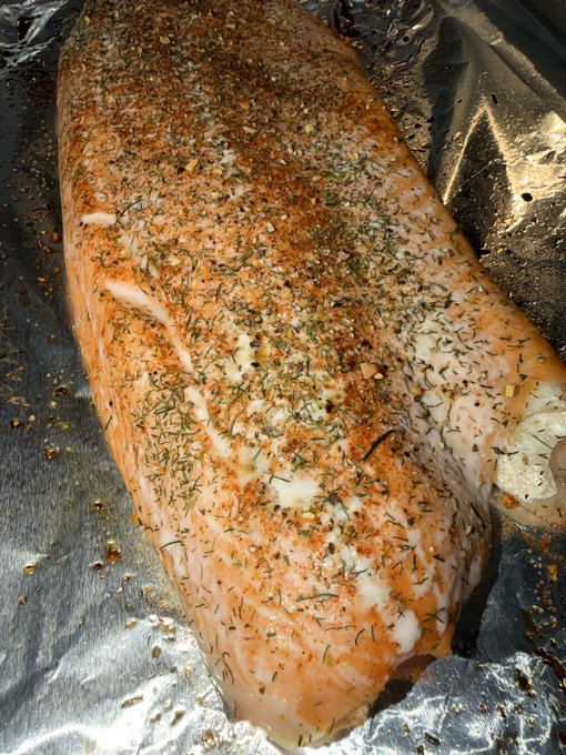 A seasoned raw fish fillet, likely salmon, covered in herbs and spices, resting on aluminum foil prepared for cooking.