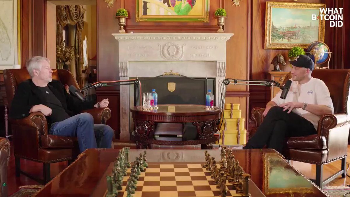 The image shows two men sitting in chairs around a table with a chess board set up between them. The room has wooden furniture and a fireplace, creating a cozy atmosphere for their conversation. One man appears to be asking a question about Bitcoin or cryptocurrency, while the other man is responding to his question.