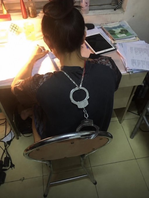 A woman with dark hair in a bun is seen from behind, writing at a desk with a tablet, wearing handcuffs around her neck attached to the chair.