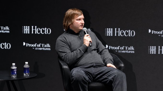 The image features a man sitting in a chair, holding a microphone and speaking into it. He appears to be at an event or conference, possibly addressing an audience. The background includes a black wall with various logos and text, including "HECTO" and "PROFOUND TALK". There are also two water bottles placed on the table next to him.