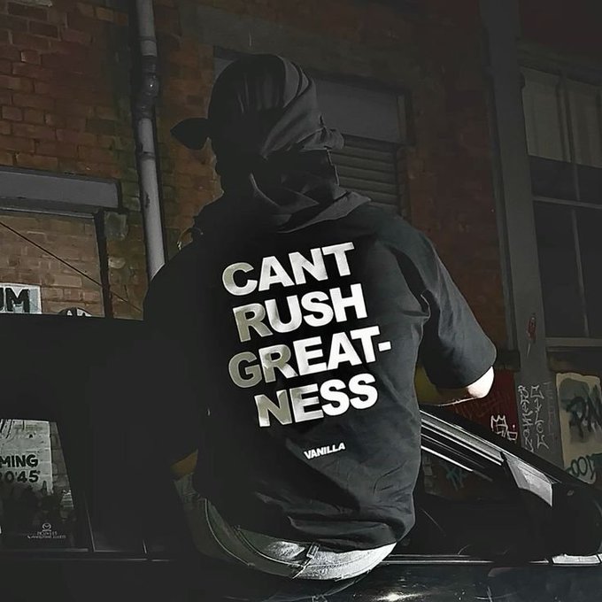 A person wearing a black hoodie with the text 'CANT RUSH GREATNESS' and 'VANILLA' printed on the back stands next to a car at night in an urban alleyway.
