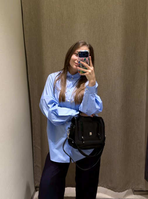 A woman with long hair takes a mirror selfie while wearing an oversized light blue button-down shirt and holding a black handbag in front of a curtain.