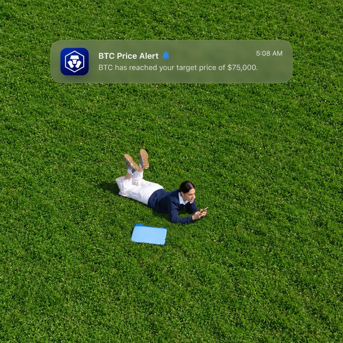 A man in business attire lying on grass while looking at his phone, with a notification above reading 'BTC Price Alert: BTC has reached your target price of $75,000' at 5:08 AM.