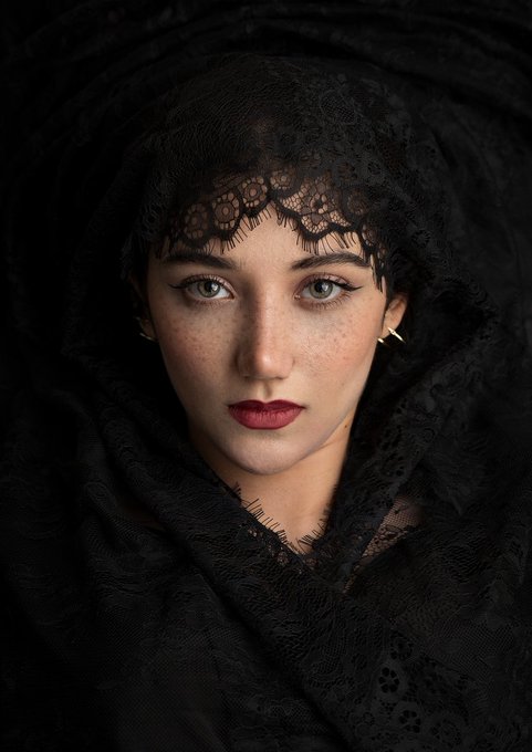 A close-up portrait of a young woman with striking light eyes and freckles, wearing dark red lipstick and a black lace veil that frames her face.