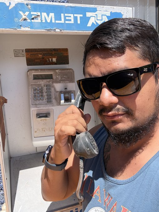 A man wearing sunglasses and a blue t-shirt speaks on the receiver of a weathered payphone with a 'TELEMEX' sign visible in the background.