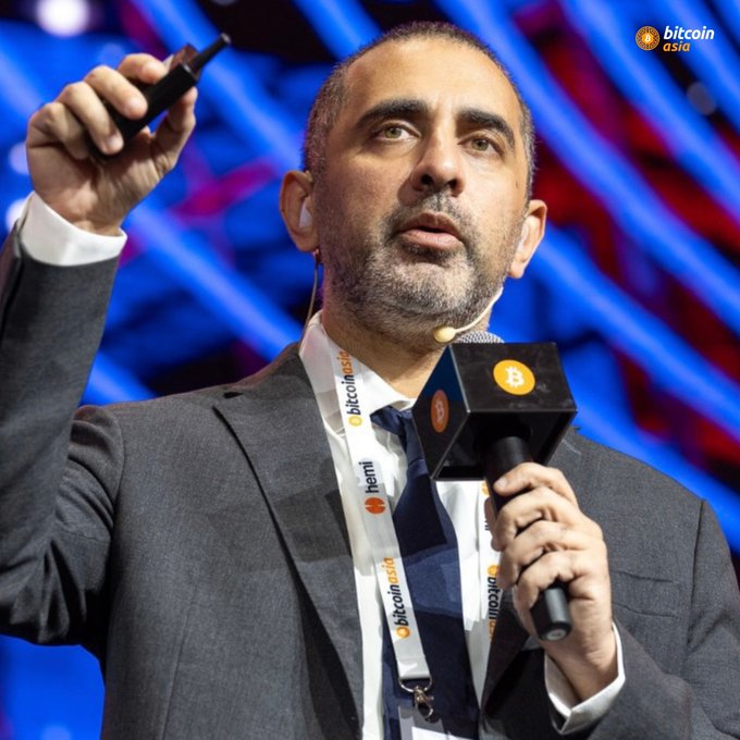 A man in a grey suit speaks into a microphone on stage at the 'Bitcoin Asia' conference, wearing a lanyard bearing the same event logo and sponsor names.