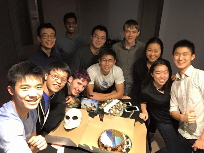 A group of young adults pose for a selfie-style photo around a table featuring a decorated cake and a white mask.