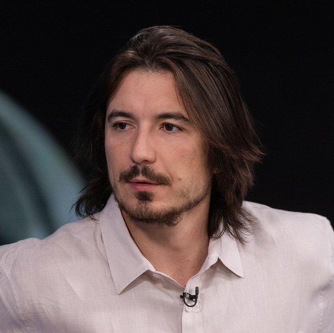 A man with shoulder-length brown hair, a mustache, and goatee, wearing a white button-up shirt with a small lapel microphone, appears to be speaking on a panel or interview setting with a dark background. The image doesn't directly depict the post about Robinhood Chain's DEX liquidity surpassing $100 million, but suggests the man may be discussing or commenting on the Robinhood-related news in a media appearance.