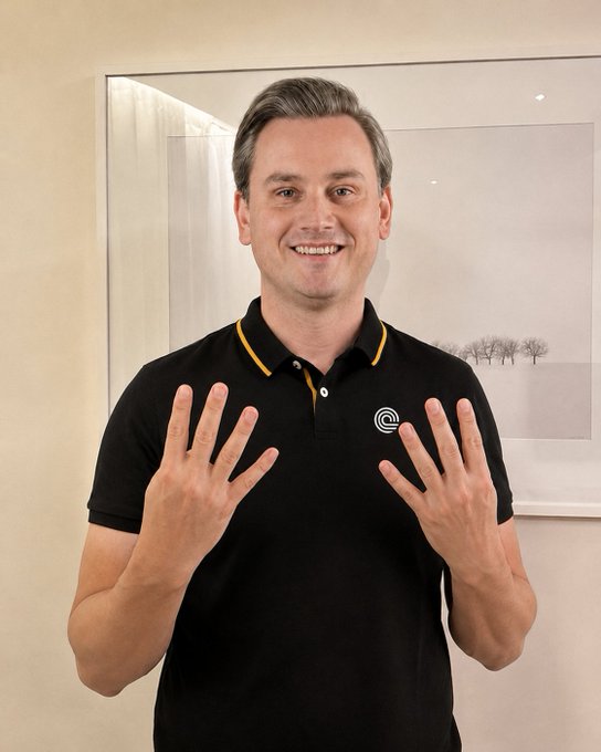A smiling man with graying hair wearing a black polo shirt with yellow trim holds up both hands, displaying all ten fingers.