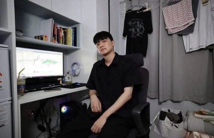 A young man wearing a black shirt sits in a gaming/office chair in front of a computer displaying a trading chart, with a bookshelf and hanging clothes visible in the background.