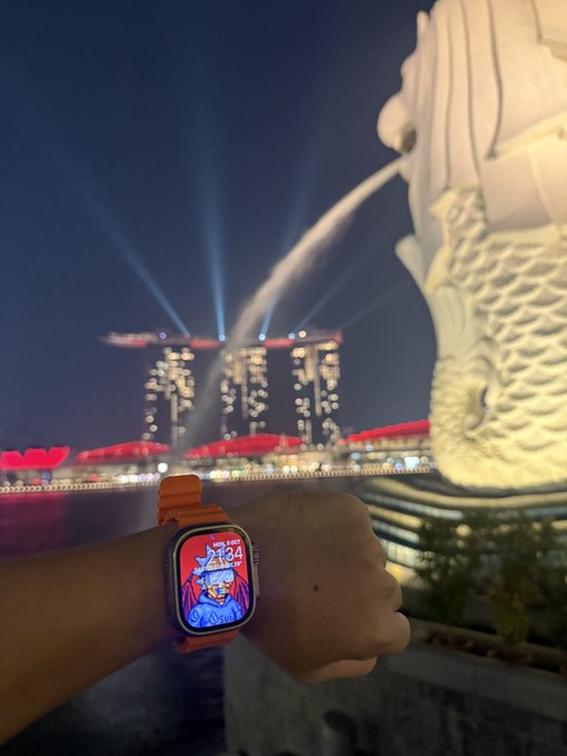 A nighttime photograph showing a wrist wearing an Apple Watch with an orange band displaying a cartoon character watch face, with the Merlion statue and Marina Bay Sands in the Singapore background.