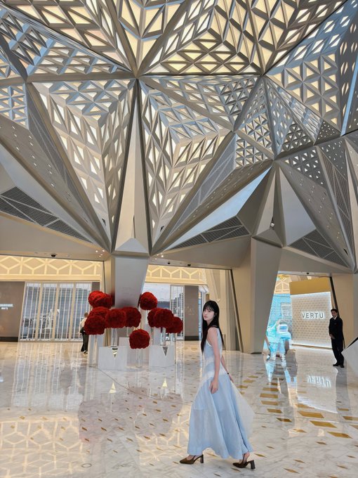 A woman in a flowing light blue dress stands in a grand, modern interior with an intricate geometric ceiling, near a 'VERTU' storefront and red floral arrangements.