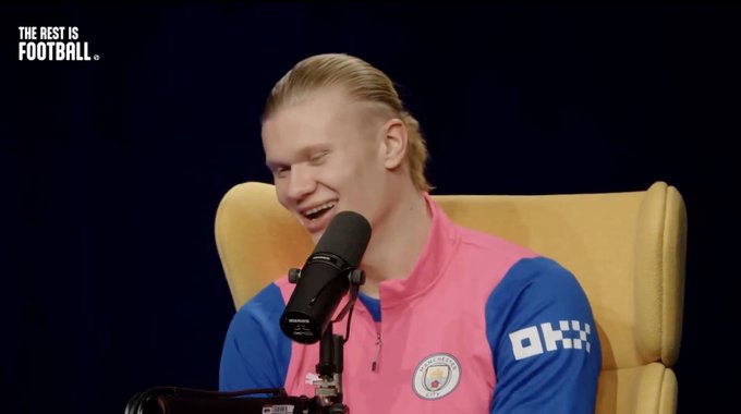 Erling Haaland, wearing a pink and blue Manchester City training top, is laughing into a microphone during a podcast, with the text 'THE REST IS FOOTBALL' visible in the corner.