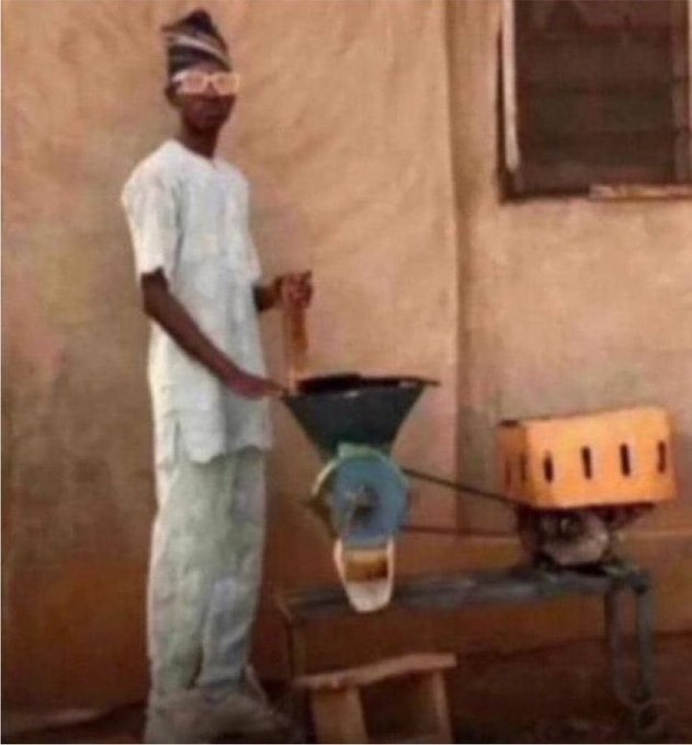 A photograph of a man wearing a hat and glasses, dressed in white traditional attire, operating a small blue grinding machine powered by a yellow motor in an outdoor setting.