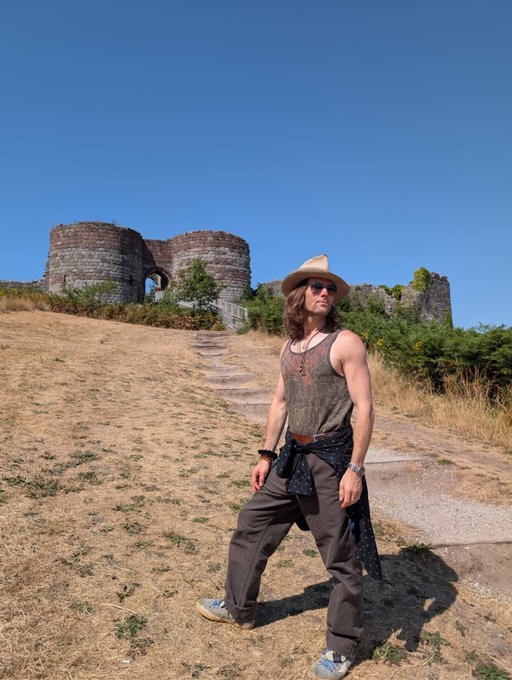 A person stands on a dry, grassy path leading up to ancient stone ruins, wearing a wide-brimmed cowboy hat, sunglasses, a patterned tank top, loose cargo pants, and a dark sash tied around the waist. The pairing with the Buddhist quote about impermanence and self-effort creates a striking visual metaphor: the weathered, partially crumbled ruins behind them embody the impermanent nature of all things, while their forward-facing stance on the path toward the ruins symbolizes the diligent, mindful journey toward inner freedom.