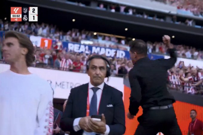 A TV broadcast showing a soccer match with the score Real Madrid 2-1 Atlético at 85:02, featuring men in suits with headsets near the field with a Real Madrid banner in the background.