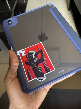 A hand holds a black tablet with a blue protective case, displaying an Apple logo and a sticker featuring a stylized robotic figure with one glowing blue eye on a red background.