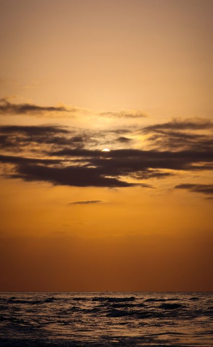 A serene sunset scene over the ocean with the sun partially obscured by dark clouds, casting a warm orange glow across the sky and reflecting on the water.