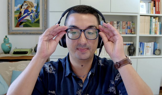 A man wearing glasses and a dark blue patterned shirt is adjusting black headphones on his head, sitting in a room with a bookshelf, a colorful painting, and a small typewriter visible behind him.