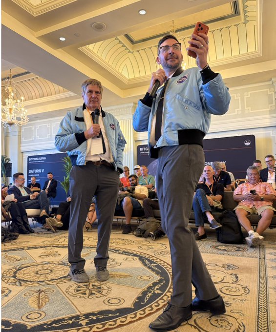 Two men in matching light blue jackets stand on a stage at the "Bitcoin Alpha 2026" event in Santa Monica, with one holding a microphone and the other filming with his phone. Visible signage displays "BITCOIN ALPHA" and "SATURDAY," aligning with the post's recap of the "Shark Tank" investor pitch sessions and thanking attendees for making the event a success.