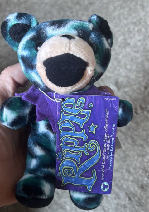 A hand holds a small plush Grimace Bear collectible by 'Liquid Death' branded with the name 'LUCKY' on its tag, sold to raise money for kids aged 3 and 14.