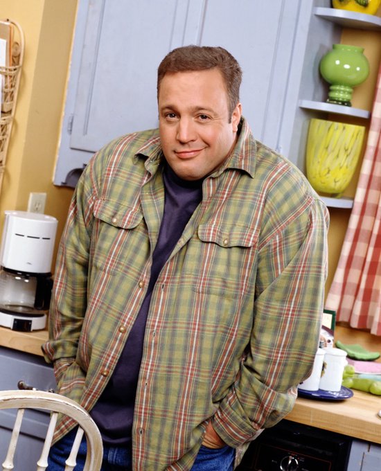A man wearing a green and red plaid shirt over a purple t-shirt, smiling in what appears to be a kitchen setting with cabinets, shelves, and a coffee maker visible.