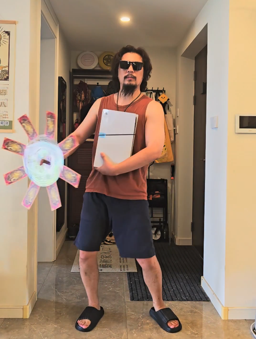 A man with long dark hair and a goatee wearing sunglasses, a brown tank top and shorts, holding a laptop and a colorful star-shaped pinwheel in a home hallway.
