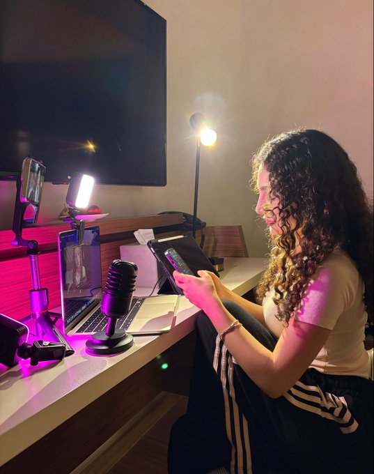 A young woman with long curly hair sits at a desk in a dimly lit room with pink accent lighting, holding a small device, with a laptop, microphone, and ring light on the desk.