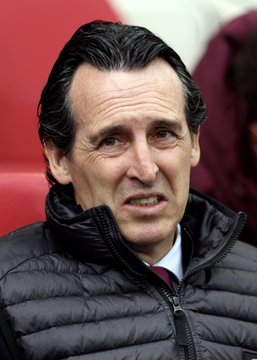 A close-up portrait of a man with dark hair, wearing a black quilted jacket over a shirt and red tie, appearing to speak with a serious expression.