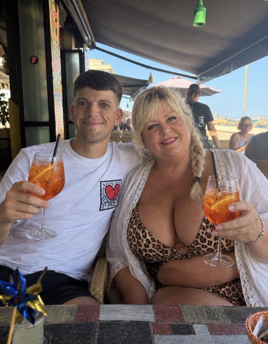 A man and woman sitting at an outdoor restaurant table clinking glasses of Aperol Spritz, with the man wearing a white 'I ❤️ YOU' t-shirt and the woman in a leopard print bikini.